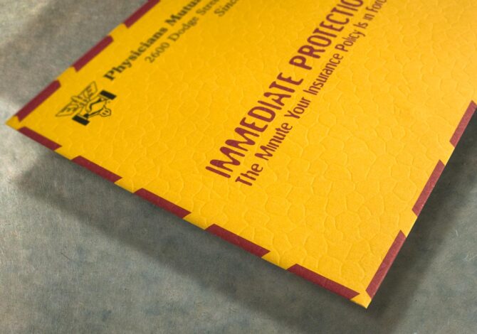 A close-up of a Tension Envelope yellow custom embossed leatherette direct mail envelope with red accents, displaying text about immediate protection insurance and a caduceus symbol in the top left corner. The envelope sits on a gray metallic surface.