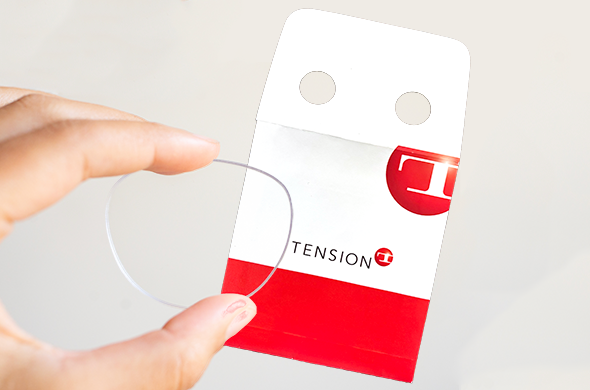 A hand holds a clear contact lens in front of a white and red optical envelope packet labeled "TENSION" with two circular holes at the top.